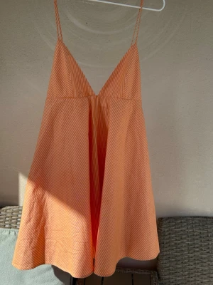  Ny trendig orange kort sommarklänning - Säljer en luftig och somrig kort klänning i orange med tunna vita ränder. Klänningen har smala axelband, v-ringning och en avslappnad passform. Perfekt för varma dagar och enkel att styla med sandaler eller sneakers.