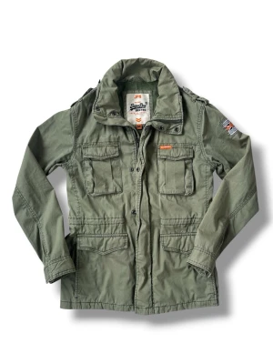 Vintage Superdry Fieldjacket Grön Storlek M - Säljer en riktigt snygg Vintage Superdry Fieldjacket som är grön storlek M. Jackan är i bra skick utan skador eller hål.  Skriv vid vidare funderingar!