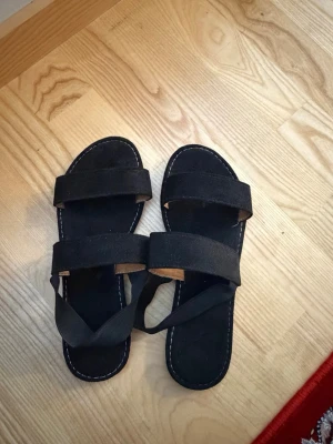 Svarta sandaler med breda band - Säljer ett par svarta sandaler med två breda band över foten och öppen tå. Sandalerna har en platt sula i brunt gummi och ovandel i mockaimitation. Snygg kontrastsöm runt sulan och enkel slip-in-modell som passar till många stilar.