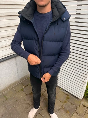 Mörkblå dunväst  - Snygg mörkblå dunväst med avtagbar huva och dragkedja framtill. Västen har en quiltad design och diskret logga på bröstet. Perfekt att slänga över en tröja för extra värme under kyliga dagar. Ny pris 4000