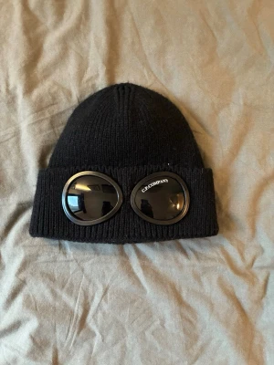 Cp company mössa - Säljer en svart ribbstickad mössa från C.P. Company med ikoniska glasögonlinser på uppviket. Mössan är i mjukt material och har en klassisk passform med bred kant. Perfekt för dig som vill sticka ut med en unik accessoar.