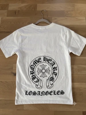 Vit Chrome Hearts t-shirt - Vit t-shirt från Chrome Hearts med stort svart tryck på ryggen och mindre logga på bröstet. Klassisk passform och rund hals. Materialet känns som mjuk bomull och trycket har tydliga detaljer med texten 'Los Angeles' på ryggen.
