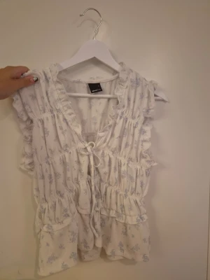 Vit blommig volangblus - Söt vit blus med ljusblått blommönster, volangdetaljer och knyt framtill. Ärmlös modell med v-ringning och avslappnad passform. Perfekt för en romantisk och somrig look. Endast använd en gång