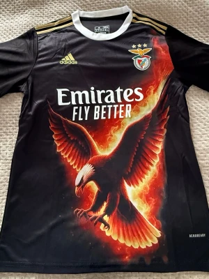 Benfica matchtröja med örntryck - Snygg svart Benfica matchtröja från Adidas med stort eldigt örntryck på framsidan, klubbmärke och tre stjärnor på bröstet samt guldfärgade detaljer på axlarna. Tillverkad i lätt och ventilerande material, perfekt för fotboll eller träning.