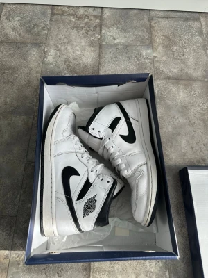 Vita och svarta Air Jordan 1  - Säljer ett par klassiska Air Jordan 1 Mid sneakers i vitt och svart läder. Skorna har den ikoniska swoosh-loggan på sidan, perforerad tåbox och svart yttersula. Perfekta för dig som gillar streetwear och vill ha en tidlös look.