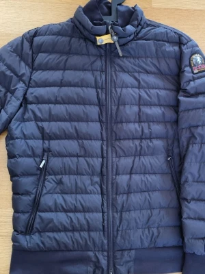 Svart pufferjacka Parajumpers Stl XL ugo - Snygg svart pufferjacka från Parajumpers med hög krage och gul detalj vid halsen. Jackan har två dragkedjeförsedda fickor och emblem på ena ärmen. Quiltad design och ribbade muddar nedtill. Perfekt för kalla dagar.