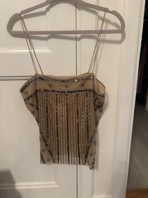 Beige mesh topp med paljetter från Zara - Trendig beige mesh topp från Zara med tunna axelband och dekorativa paljetter i svart och silver. Toppen har ett transparent tyg och ett unikt mönster framtill som ger en cool och festlig look. Perfekt att styla med jeans eller kjol för en snygg outfit.
