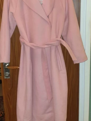 Ljusrosa kappa med knytskärp - Snygg ljusrosa kappa med klassisk krage och knytskärp i midjan. Kappan har raka ärmar, två fickor framtill och är tillverkad i ett mjukt tyg som ger en elegant look. Perfekt för dig som vill ha en stilren och trendig ytterjacka.
