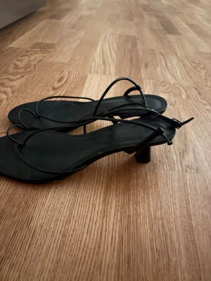 Svarta strappy sandaler med klack - Säljer ett par svarta sandaler från H&M med tunna remmar och låg klack. Sandalerna har en minimalistisk design med fyrkantig tå och justerbar rem runt ankeln. Perfekta för dig som gillar stilrena och trendiga skor.