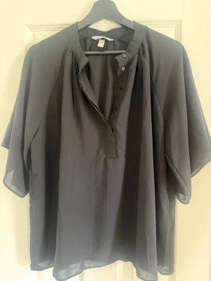 Svart blus från H&M med knappar - Stilren svart blus från H&M med lös passform och korta ärmar. Blusen har knappar framtill vid halsen och är tillverkad i ett lätt, tunt material som ger en luftig känsla. Perfekt att matcha med jeans eller kjol för en trendig look.