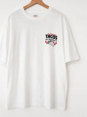 Ny Vit oversized t-shirt med taco-tryck - Vit oversized t-shirt från ASOS med färgglatt taco-inspirerat tryck fram och stort tryck på ryggen. Texten 'El Camino TACOS Let's Taco 'Bout It!' och rutigt mönster ger en lekfull vibe. Tillverkad i mjuk bomull med rund hals och avslappnad passform.