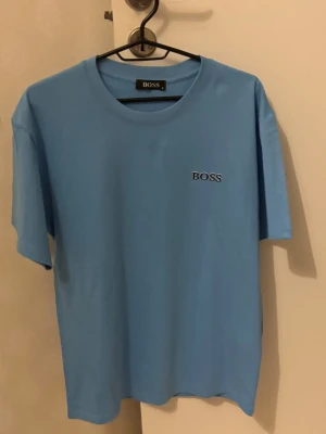 Blå BOSS t-shirt i bomull - Snygg ljusblå t-shirt från BOSS med klassisk rund hals och diskret BOSS-logga på bröstet. Tillverkad i mjuk bomull för en skön känsla. Perfekt till jeans eller shorts för en clean och stilren look.