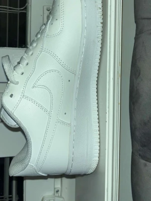 Nike Air Force 1 vita sneakers - Klassiska helvita Nike Air Force 1 sneakers i lågt utförande. Skorna har en stilren design med Nike swoosh på sidan och AIR-broderi på hälen. Tillverkade i slätt läder med matchande vita skosnören och sula. Perfekta för en clean och tidlös look.