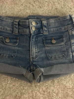 Blå jeansshorts med fickor - Säljer ett par blå jeansshorts med dubbla knappar framtill och två framfickor med lock och knapp. Finns klippta från ett par jeans 