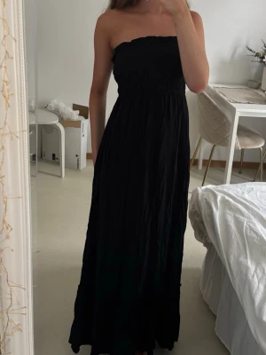 Svart axelbandslös långklänning - Svart maxiklänning i bandeau design och resår i midjan. Klänningen har en enkel och stilren look med lätt utställd kjol. Perfekt för sommarens varma dagar eller när du vill ha en avslappnad men snygg outfit.