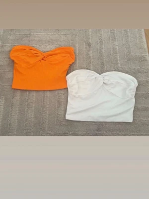 Två bandeau tubtoppar i orange och vitt - Säljer två bandeau tubtoppar, en i stark orange och en i vitt. Båda har twistad detalj framtill och är ribbade i materialet. Perfekta för sommaren och passar till både jeans och kjol.