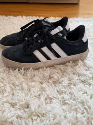 Marinblåa Adidas sneakers med vita ränder - Klassiska marinblåa Adidas sneakers med vita detaljer och de ikoniska tre ränderna på sidan. Skorna har marinblå ovandel i mockaliknande material, vita sulor och vita hälpartier. Låga i modellen och med marinblåa skosnören. Perfekta för en avslappnad streetstyle.