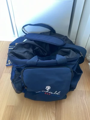 Mörkblå ryktarväska Fairfield - Rymlig mörkblå ryktarväska från Fairfield Sports Design med broderad logga och hästmotiv. Väskan har flera fack, justerbara axelband, bärhandtag och nätficka på sidan. Tillverkad i slitstarkt tyg, perfekt för stall och ridning. Lite smått och gott med ryktarsaker includeras.