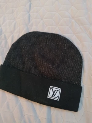 Louis Vuitton mössa, svart - Snygg svart mössa från Louis Vuitton med uppvikt kant och klassisk LV-logga framtill. Tillverkad i ett mjukt, stickat material som känns lyxigt och passar perfekt till höst och vinter. Enkel och stilren design som funkar till alla outfits. Den är varsamt använd skriv gärna till mig om du har  frågor!