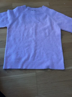 Ljuslila stickad tröja - Mysig ljuslila stickad tröja med rund hals och ribbade muddar vid ärmslut och nederkant. Tröjan har en enkel och avslappnad passform, perfekt för att hålla värmen. Materialet ser ut att vara mjukt och bekvämt, troligen bomull eller en bomullsblandning.