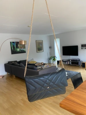 Svart quiltad axelväska med kedja - Snygg svart axelväska i quiltat mönster med guldfärgad kedjerem. Väskan har en stilren design med viklock och liten metallogo framtill. Perfekt för dig som vill ha en trendig och praktisk väska till alla tillfällen.
