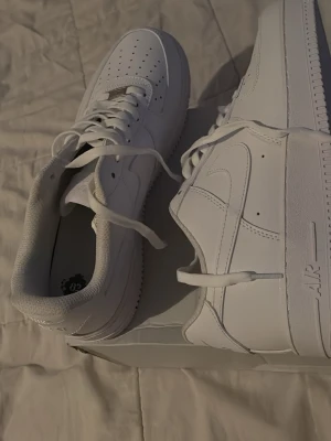 Nike Air Force 1 vita sneakers - Klassiska helvita Nike Air Force 1 sneakers i lågt utförande. Skorna har en stilren design med perforerad tå, kraftig sula och Nike swoosh på sidan. Tillverkade i slätt läder med vita skosnören och detaljer. Perfekta för dig som gillar tidlös streetstyle.