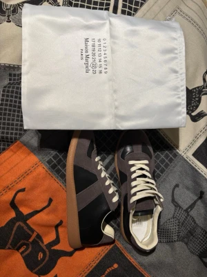 Maison Margiela sneakers svart/brun - Maison Margiela sneakers i svart och mörkbrunt läder och mocka med vita snören och gummisula. Klassisk low-top siluett och diskret branding på plösen. Snygg retrostil som passar till många outfits. Dem är använda typ 2-3 gånger 