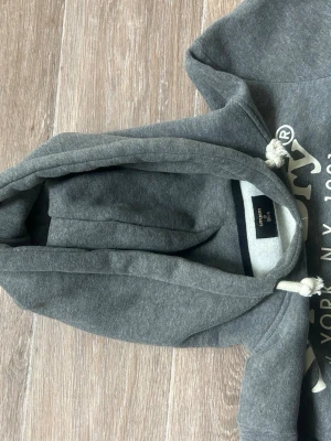 Grå hoodie från Superdry med tryck - Snygg grå hoodie från Superdry med vitt tryck på bröstet och textdetaljer. Klassisk känguruficka framtill och justerbar huva med vita snören. Perfekt för en avslappnad streetwear-look.