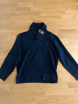 Polo Ralph Lauren - Stickad tröja från Polo Ralph Lauren med sjalkrage och knappdetaljer vid halsen. Tröjan har lång ärm och den klassiska gröna logotypen broderad på bröstet. Perfekt för dig som gillar stilrena och tidlösa plagg.