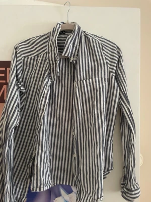 Randig skjorta från Rut&Circle XS - Snygg skjorta från Rut&Circle i storlek XS. Skjortan har vertikala blå och vita ränder, klassisk krage, bröstficka och knäppning framtill. Tillverkad i ett lätt och luftigt material som passar perfekt till varmare dagar.