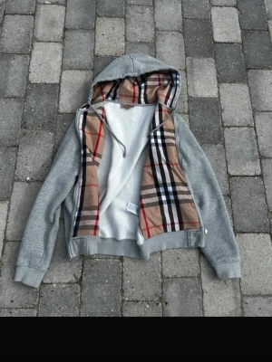 Grå hoodie med rutigt foder - Snygg grå hoodie med dragkedja och huva. Insidan av huvan och öppningen har ett klassiskt beige, svart, vitt och rött rutmönster. Mjuk fleece på insidan för extra komfort. Perfekt för dig som vill ha en stilren men ändå unik hoodie.