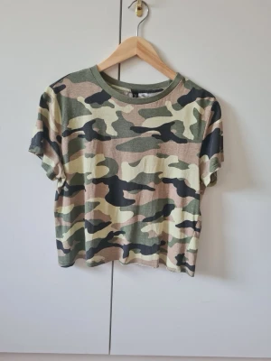 Camouflage topp med kort ärm från H&M strl M - Säljer en kortärmad topp från H&M med klassiskt camouflage-mönster i grönt, beige och svart. Material 100% bomull. Storlek M.