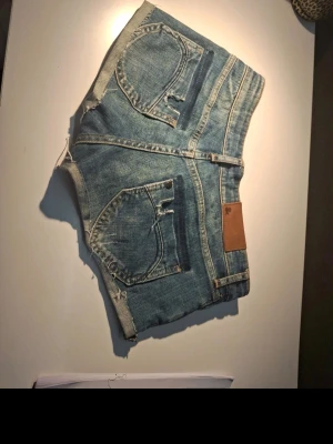 Blå jeansshorts med rå kant - Säljer ett par blå jeansshorts med råa kanter och klassiska fem fickor. Shortsen har bälteshällor, knappstängning och en brun patch bak i midjan. Perfekta för varma dagar och har en avslappnad vibe.