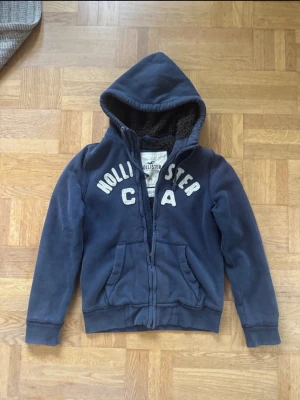 Blå hoodie från Hollister med dragkedja - Mysig blå hoodie från Hollister med dragkedja och broderad text på bröstet. Fodrad huva med mjukt teddyfoder och två stora fickor framtill. Perfekt för chill dagar och enkel att matcha med jeans eller mjukisbyxor.