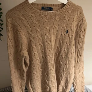 Beige kabelstickad tröja från Ralph Lauren - Säljer en klassisk beige kabelstickad tröja från Ralph Lauren med rund halsringning. Tröjan har en diskret logga broderad på bröstet och långa ärmar. Perfekt för kyligare dagar.
