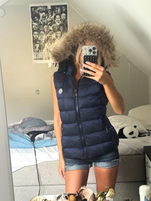 Marinblå Moncler väst med pälshuva - Snygg skimmerblå dunväst från Moncler med äkta pälskrage. Västen har två dragkedjefickor, varav höger fickan inte går att stänga. Annars toppen skick🩵storlek 1 us, passar storlek xs/s ! Jag är 167 cm lång