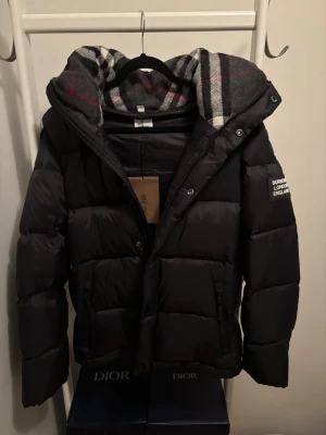 Svart pufferjacka från Burberry - Snygg svart pufferjacka från Burberry med patch på ärmen där det står 'Burberry London England'. Jackan har en stor huva med klassiskt rutigt foder i grått, vitt och rött. Dragkedja framtill och två fickor med dragkedja. Perfekt för kalla vinterdagar.