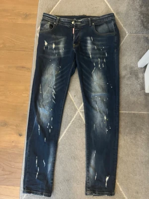 Mörkblå jeans med färgstänk - Säljer ett par mörkblå jeans med coola slitningar och vita färgstänk på både fram- och baksida. Jeansen har klassisk femficksmodell och smal passform. Perfekta för dig som gillar en trendig och avslappnad streetstil.