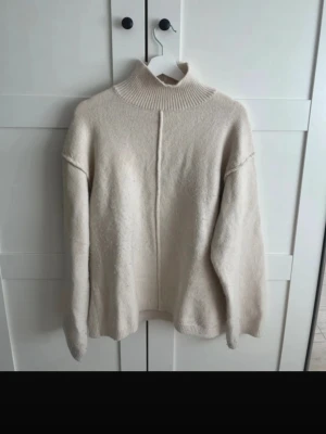 Beige stickad polotröja från H&M - Mysig beige stickad polotröja från H&M med rak passform och markerad söm framtill. Tröjan har ribbad hög krage och långa ärmar, perfekt för kyliga dagar. Enkel och stilren design som är lätt att matcha med olika outfits.