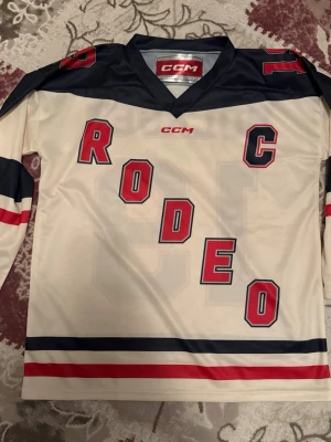 Vit och marinblå hockeytröja CCM - Vit långärmad hockeytröja från CCM med marinblå och röda detaljer. Framsidan har texten RODEO och C, baksidan har BOLAGET och siffran 19. Tröjan har breda ränder på ärmar och nederkant och är gjord i ett sportigt syntetmaterial.
