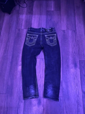 Svarta True Religion jeans - Säljer ett par svarta jeans från True Religion med tydliga vita kontrastsömmar och klassiska bakfickor med U-formad design. Modellen är Joey Super T och har en avslappnad passform. Perfekta för dig som gillar streetwear och vill sticka ut.