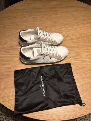 Vita Philippe Model sneakers med blå detalj - Snygga vita sneakers från Philippe Model med beige mockadetaljer och mörkblå häl. Klassisk låg modell med vita skosnören och ikonisk sköld på sidan. Skorna är i läder och mocka, perfekta för en clean och stilren look.