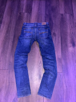 Blåa jeans från nudie - Säljer ett par mörkblå jeans med raka ben och klassiska fem fickor. Jeansen har snygga slitningar och kontrastsömmar. Dragkedjegylf och bälteshällor. Materialet är denim i bomull. Perfekta för en avslappnad och trendig look.