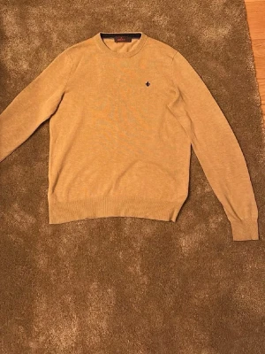 Beige stickad Morris tröja  - En stilren beige stickad tröja med rund hals och ribbade muddar vid ärmslut och nederkant. Tröjan har en liten mörkblå broderad detalj på bröstet. Perfekt för lager-på-lager och enkel att matcha med olika outfits.