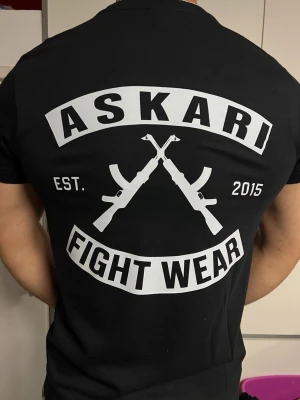 ASKARI Fight Wear svart T-shirt - Svart ASKARI T-shirt med stilrent tryck framtill och stort “Fight Wear”-tryck på ryggen. Snygg och bekväm passform. I mycket fint skick.