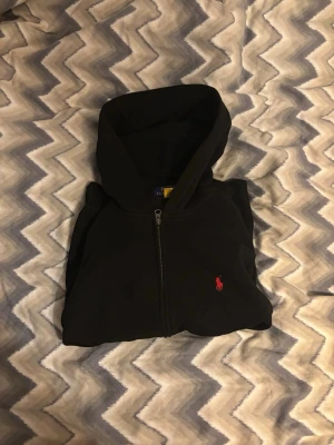 Äkta Ralph lauren hoodie - Säljer en svart Ralph lauren hoodie med dragkedja och huva. Perfekt för chill dagar eller när du vill ha en enkel och stilren look. Storlek 160/ 14-16 år. Mycket bra skick men ett litet jack som inte märks av