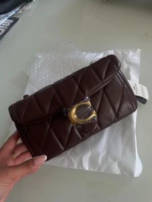 Mörkbrun Coach handväska - Snygg mörkbrun quiltad handväska i läder med guldfärgade detaljer och kedjerem. Väskan har ett stort C-spänne framtill och en klassisk rektangulär form. Perfekt accessoar för att lyfta din outfit.