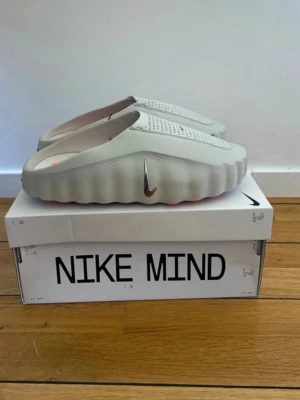 Nike Mind grå tofflor - Säljer ett par Nike Mind tofflor i ljusgrått med vågig sula och ventilerande hål på ovansidan. Tofflorna har en futuristisk design och en liten silverfärgad Swoosh på sidan. Perfekta för chill dagar hemma eller när du vill sticka ut.