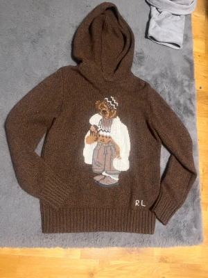 Polo Ralph lauren hoodie  - Mysig brun stickad hoodie med en stor broderad björn på framsidan. Tröjan har huva, ribbade muddar och RL-broderi nedtill. Perfekt för dig som gillar unika detaljer och streetstyle-vibe. Har ägt denna sköna hoodie ett tag nu och är sugen på något nytt. Har knappt använt den för att den ör för liten för mig 