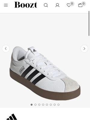 Vita Adidas sneakers - i ny förpackning  - Klassiska vita sneakers från Adidas med svarta ränder på sidan och brun sula. Skon har en låg profil, vit ovandel i läder och mockadetalj vid tån. Perfekt för dig som gillar stilrena och sportiga skor. I ny förpackning och aldrig använda 